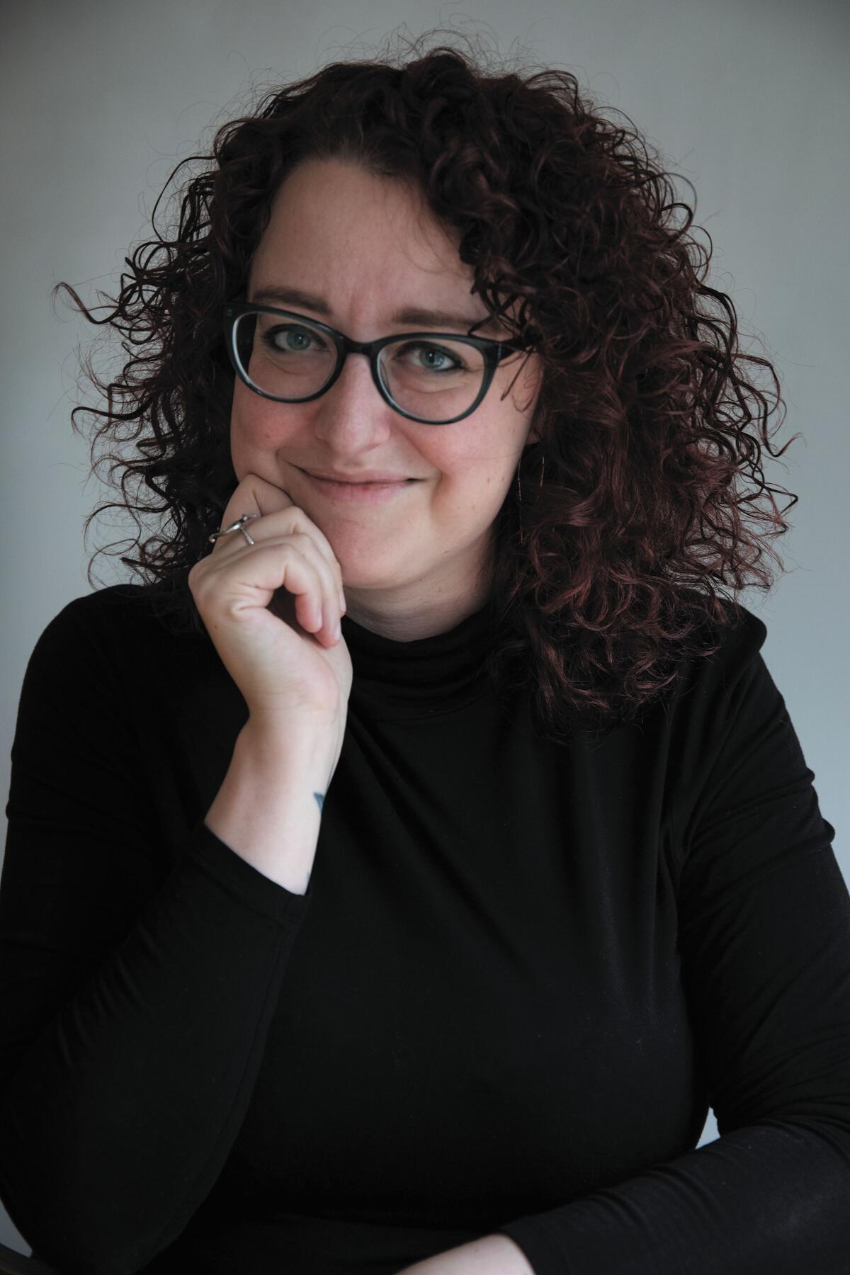 jeanna kadlec author headshot; a white woman with red curly hair and blue glasses smiling while wearing a black turtleneck and silver rings