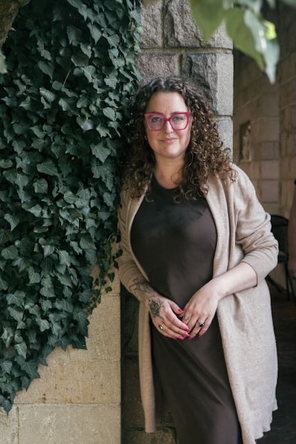 curly haired white woman with pink glasses wearing a silk brown dress and oatmeal cashmere cardigan next to crawling ivy